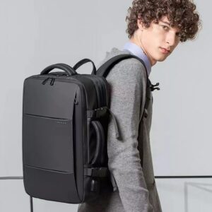 Un jeune homme portant à l'épaule un sac à dos valise rectangulaire noir. Celui-ci se ferme avec une glissière sur le dessus et a une hanse sur ses deux faces.